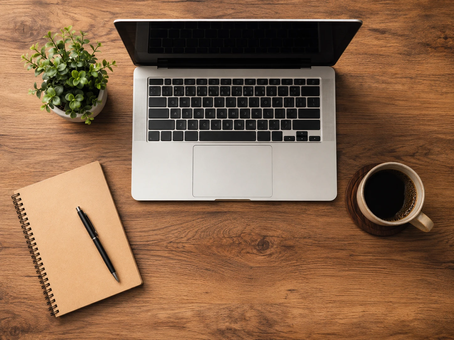 Clean wooden desk workspace with a laptop, plant, notebook, pen, and coffee